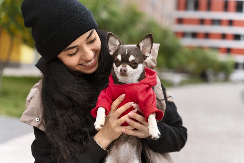 mujer con su chihuahua en brazo con polerón rojo ambos en exterior y abrigados en un día de invierno