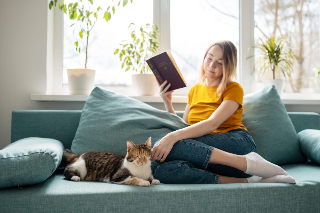 tutora acaricia mientras mira a su gato ambos estan sobre el sofa con luz fia y ella tiene un libro en su mano